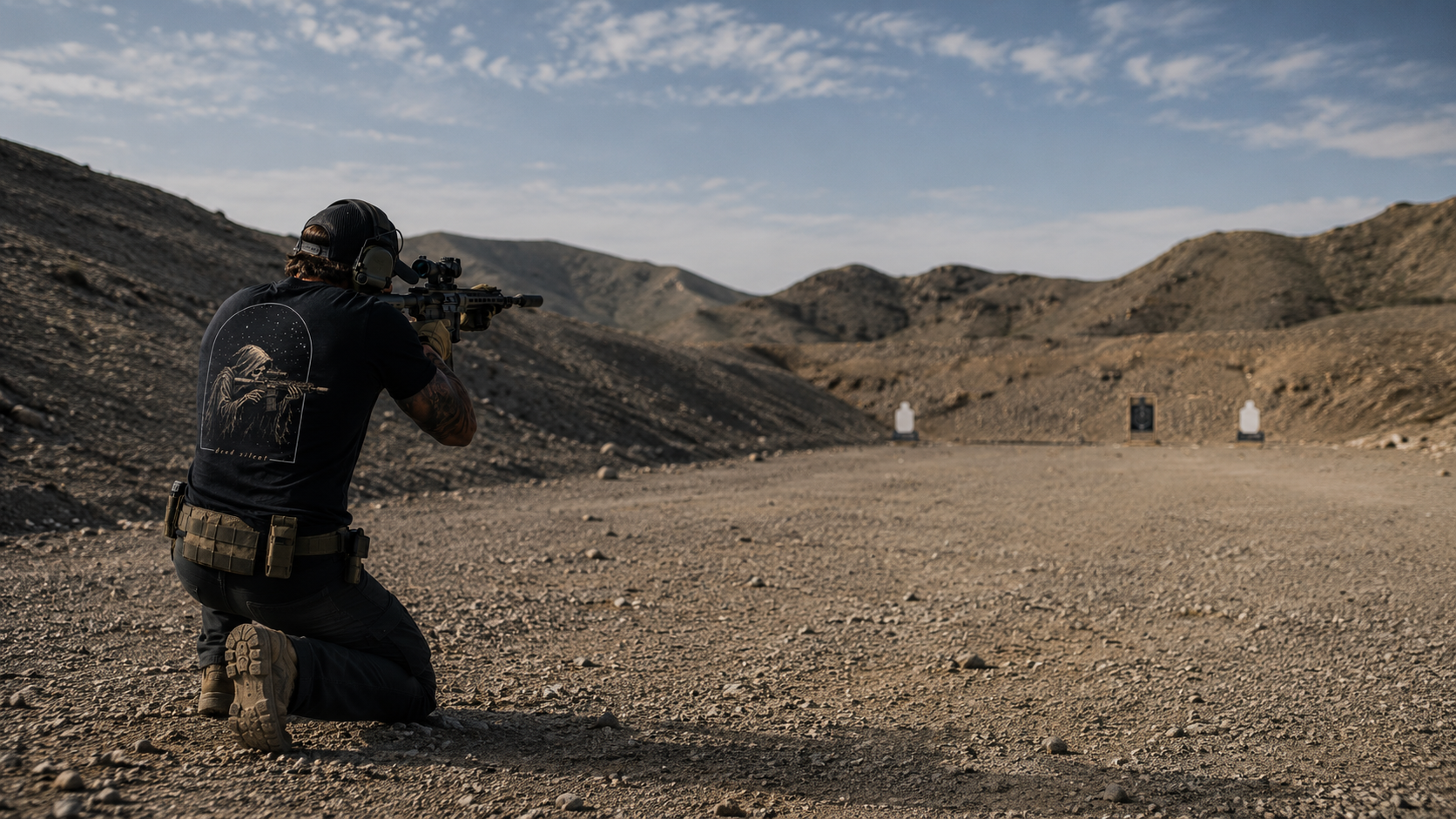 Shooter kneeling on a desert range wearing an Outlaw Standard tee